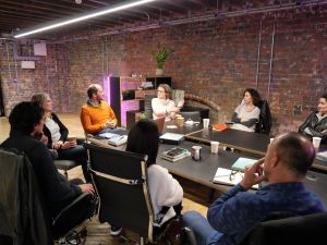 A group of 8 adults sat around a table in an office space looking at 2 guest speakers. With note books and coffee.