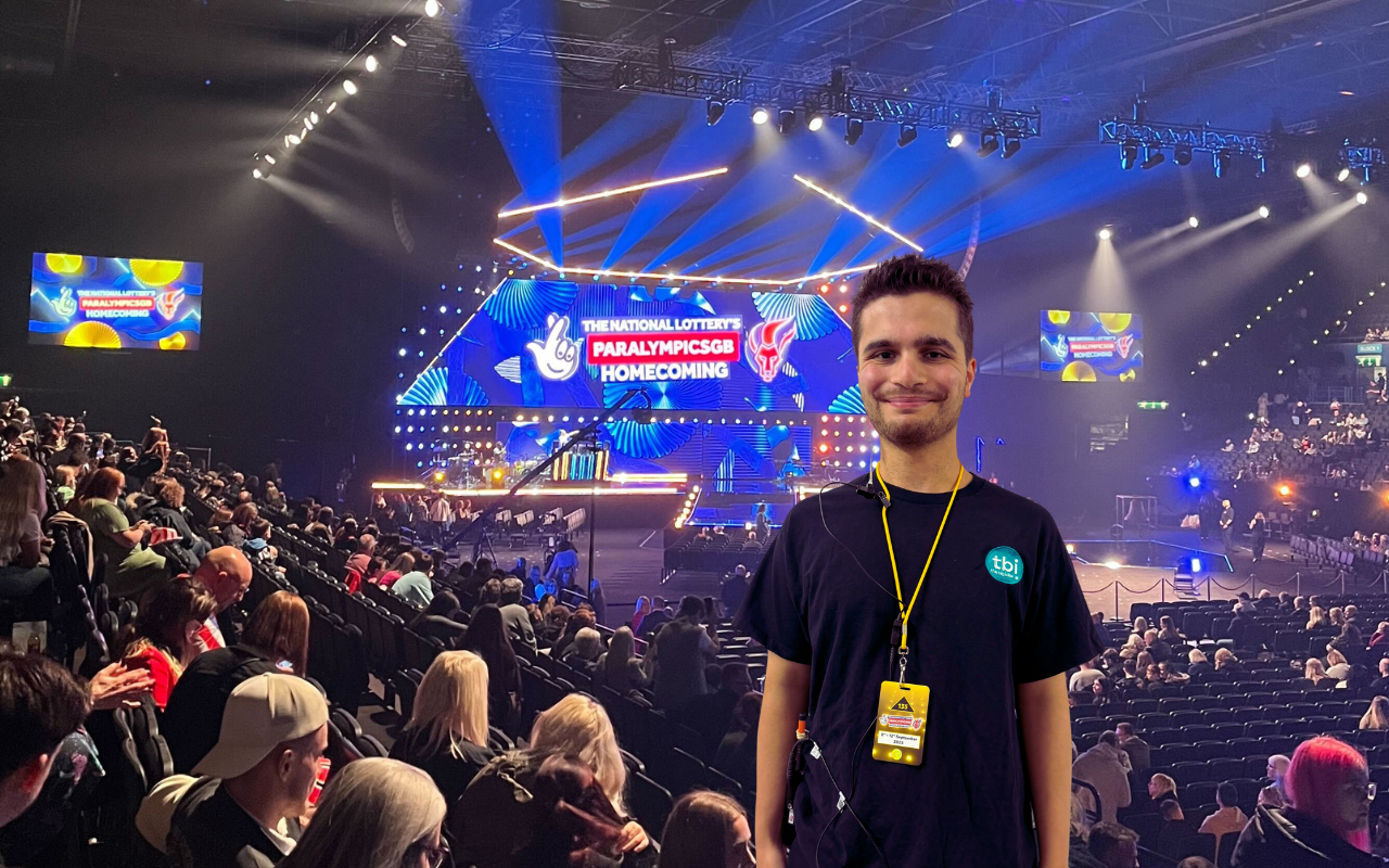 Luca standing in front of the ParalympicsGB Homecoming Ceremony stage at the Utilita Arena in Birmingham. He is wearing a black T-shirt with the ‘tbi’ logo and a yellow lanyard. The background shows a vibrant stage setup with lights, a large screen displaying the event name, and a seated audience