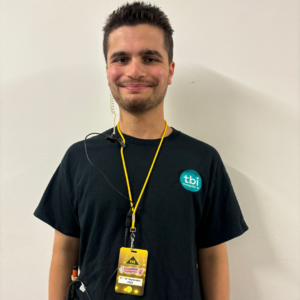 Luca standing against a plain white wall, wearing a black T-shirt with the ‘tbi’ logo and a yellow lanyard with his event pass for the Paralympics Homecoming Ceremony. He has an earpiece in his ear, smiling confidently.